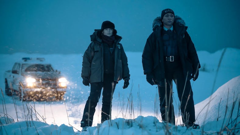 Two individuals in winter clothing stand in the snow, with a car's headlights shining in the backgro...