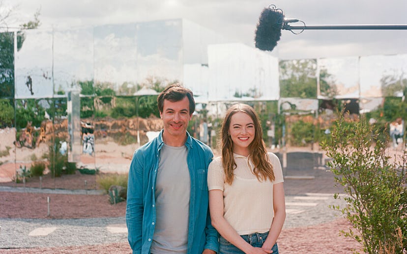 Two smiling people standing in front of mirrored surfaces outdoors with a microphone boom overhead.