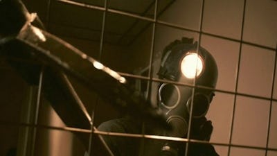Person wearing a gas mask with a light on the side behind a grid, in a dimly lit tiled room.