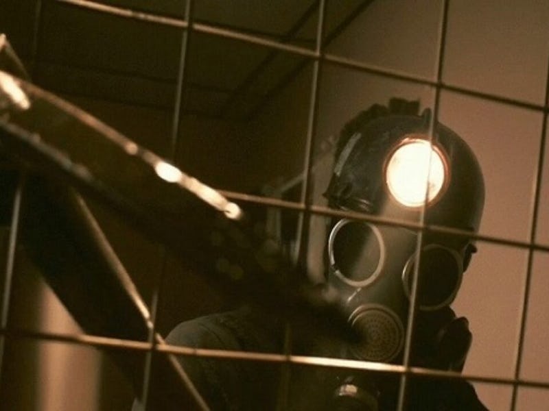 Person wearing a gas mask with a light on the side behind a grid, in a dimly lit tiled room.