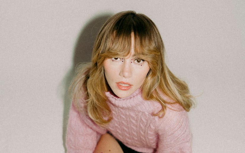Woman in pink sweater and black shorts posing with one knee bent, against a plain background.