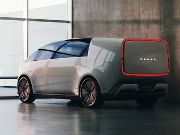 A futuristic electric vehicle with a boxy design and a red illuminated rear panel is parked in a mod...