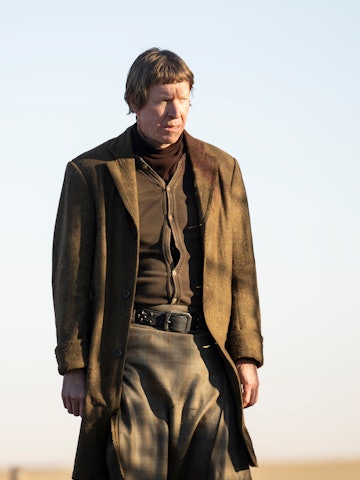 Man in a brown coat and trousers with a troubled expression standing outdoors.