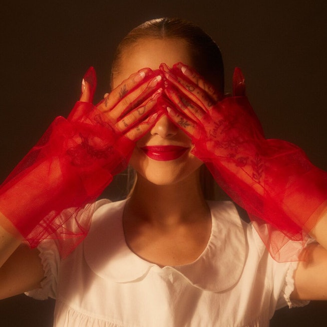 A woman with red lipstick covers her eyes with her red-stained hands wearing sheer red gloves agains...