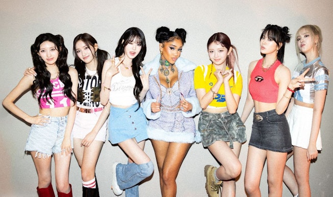 Six women from a music group and a featured artist pose together, sporting trendy outfits with a mix...