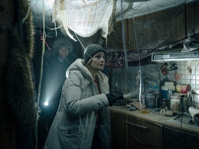 Two people in warm clothing look tense inside a dimly lit, cluttered room with hanging nets and rust...