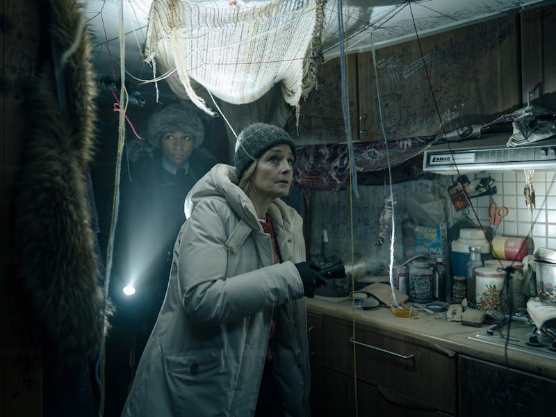 Two people in warm clothing look tense inside a dimly lit, cluttered room with hanging nets and rust...