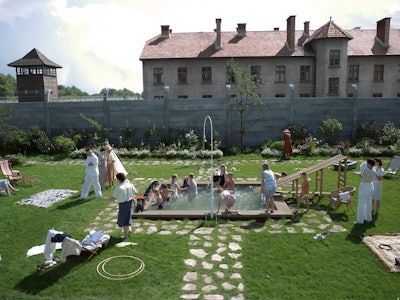 People relax in a lush courtyard with a pool, behind a sign on a building reads "Arbeit Macht Frei."