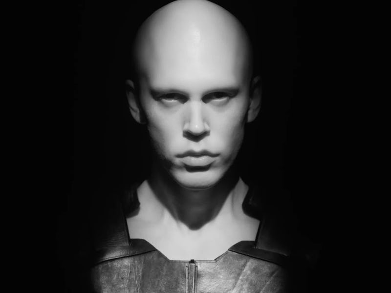 A bald man with a serious expression, emerging from shadows in a black and white photo.