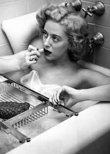 a black and white photo of a blonde person in a bathtub applying lipstick in front of a mirror that'...