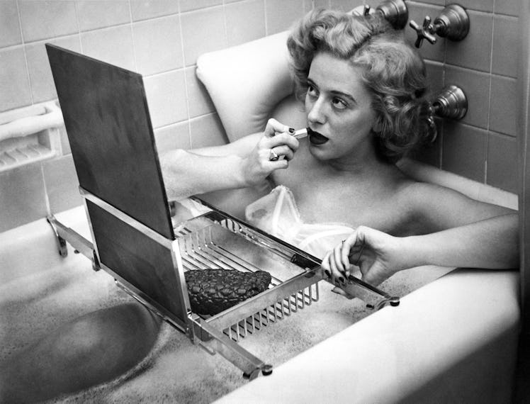 a black and white photo of a blonde person in a bathtub applying lipstick in front of a mirror that'...