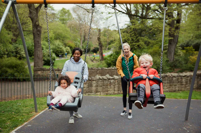 Two mom friends push their kids on the swings at the park.