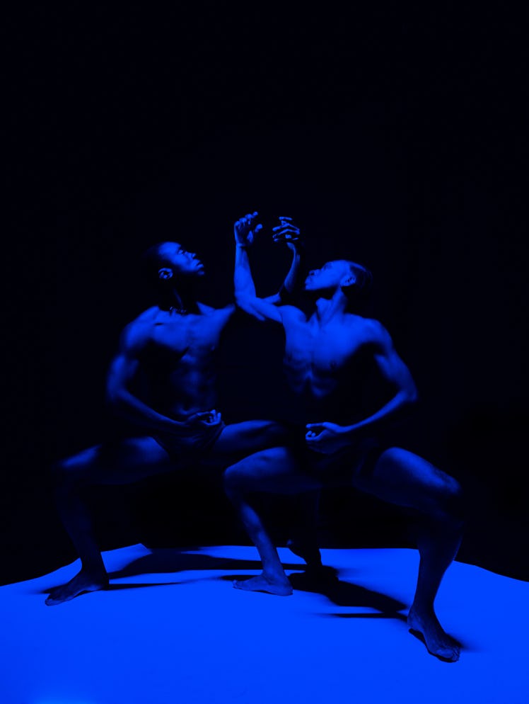 two people bathed in blue light pose in a squatting position, holding up their arms together to crea...