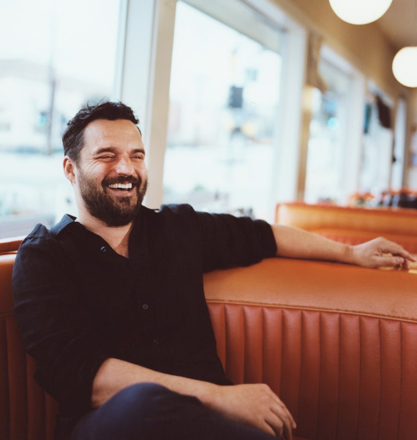 Jake Johnson laughing while sitting on an orange diner booth with windows in the background.