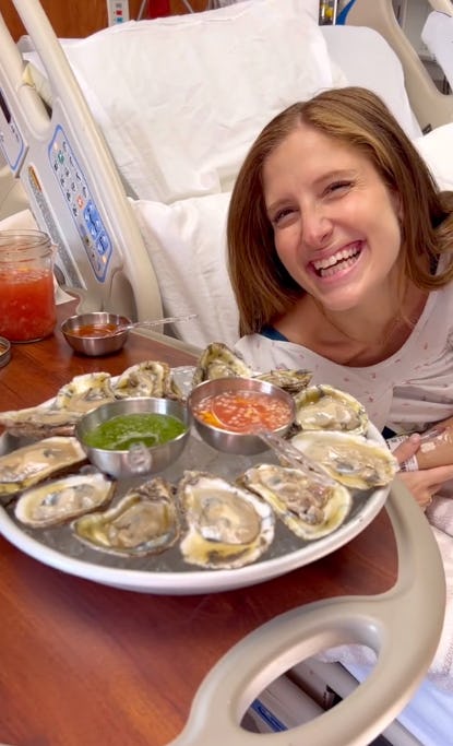 A smiling woman in a hospital bed enjoys a platter of oysters with dipping sauces, surrounded by hospital equipment and bedding.