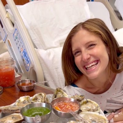 A smiling woman in a hospital bed enjoys a platter of oysters with dipping sauces, surrounded by hospital equipment and bedding.