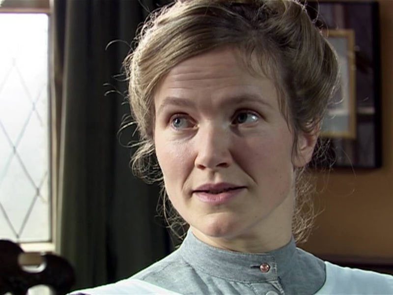 A woman with light brown hair styled in a bun looks intently at a man. She wears a gray dress with a...