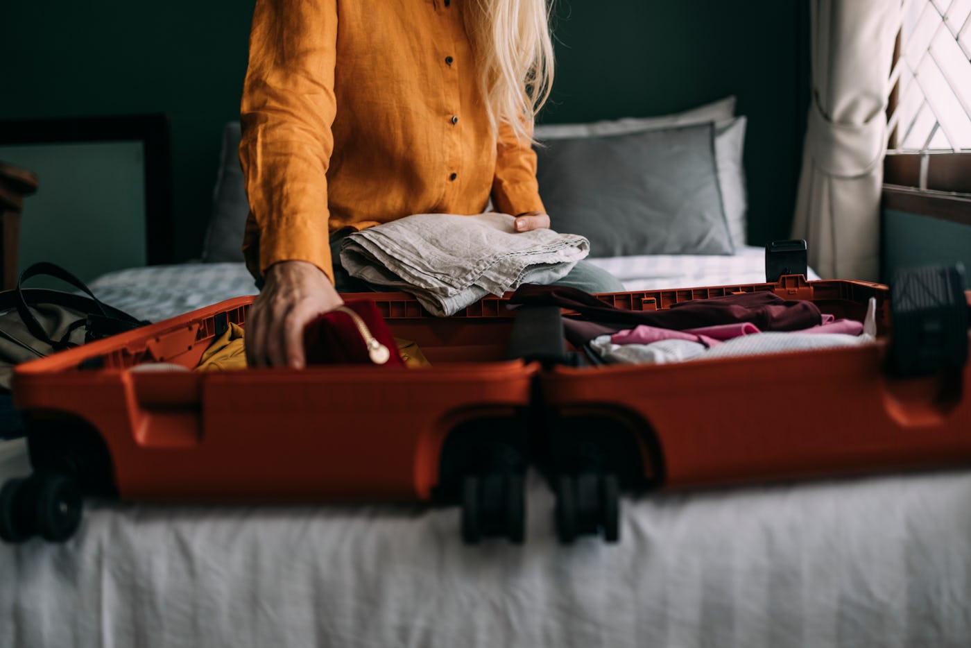 A person with long hair is packing an orange suitcase with folded clothes on a bed, surrounded by pillows and a calm, green-hued room.