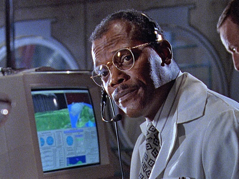 A focused man in a lab coat and glasses speaks to two colleagues in a scientific setting, with a com...