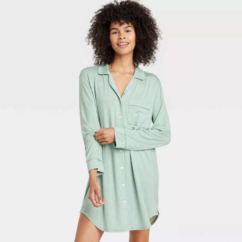 A woman wearing a light green, striped button-up shirt dress smiles while posing with one hand on he...