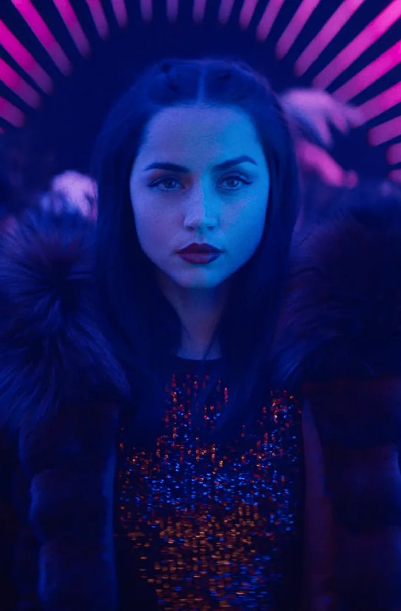 A person with long dark hair and bold lipstick stands confidently in a dimly lit space, surrounded b...