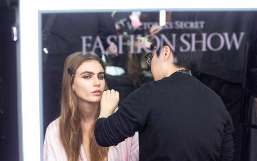 A model with long brown hair sits in front of a mirror, receiving makeup application from an artist ...