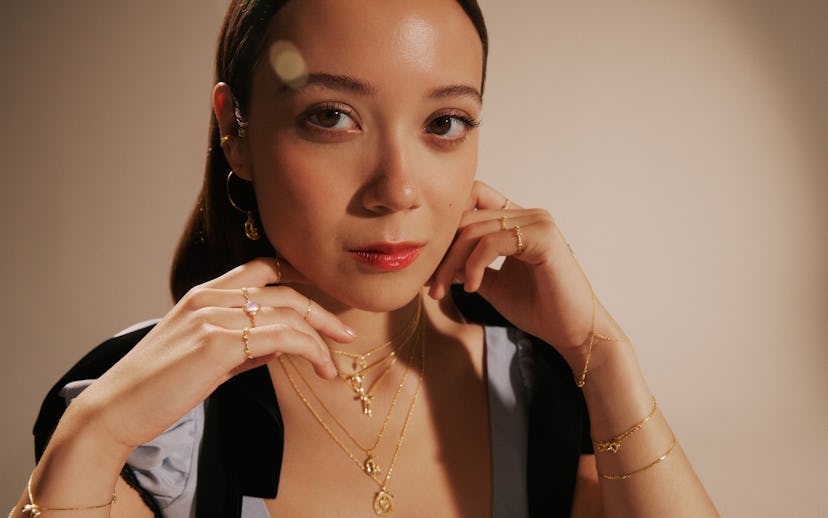 A woman with long brown hair poses elegantly, wearing multiple gold necklaces. She touches her jewel...