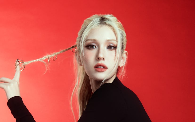 A young woman with long blond hair poses against a red backdrop, wearing a black crop top and red pa...