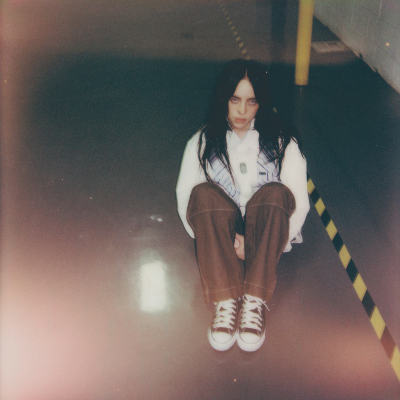 A person with long dark hair sits on the floor in a dimly lit space, wearing a white shirt, brown pa...