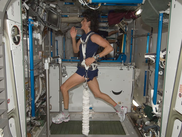 A woman in workout gear runs on a treadmill in a spacecraft, secured by harnesses, with equipment an...