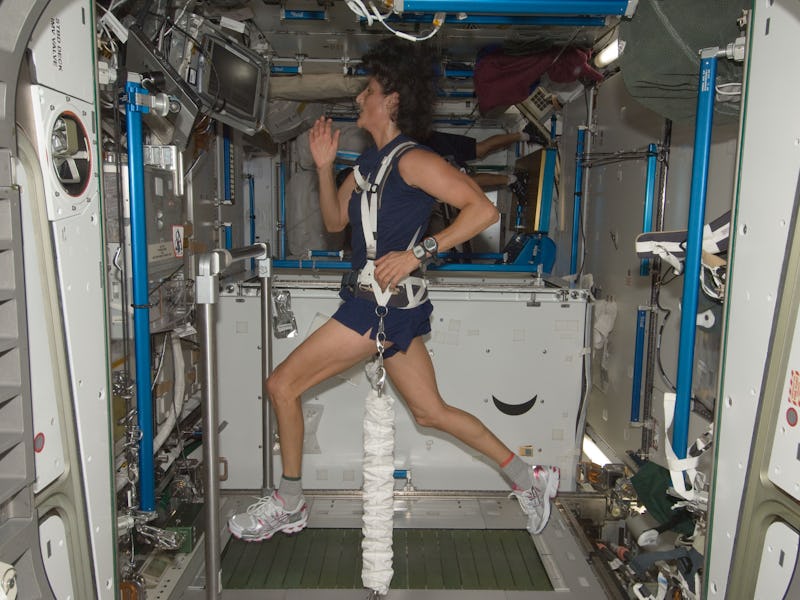 A woman in workout gear runs on a treadmill in a spacecraft, secured by harnesses, with equipment an...