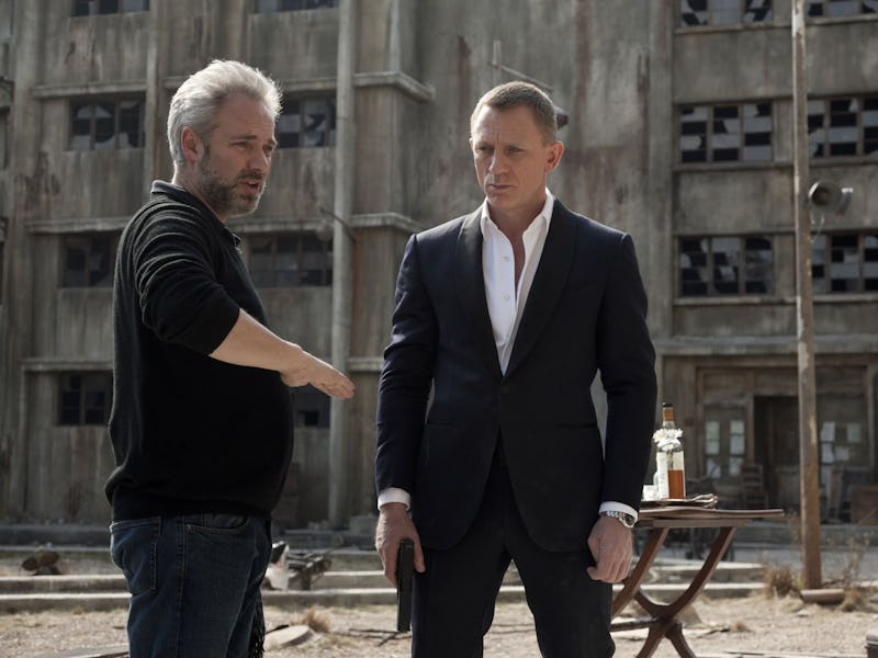 A director discusses a scene with a suited actor on an outdoor set, featuring a dilapidated building...