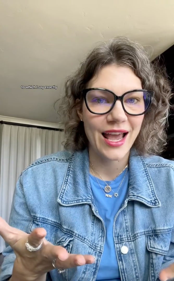 A woman with curly hair and glasses is animatedly speaking, wearing a denim jacket over a blue shirt. She gestures with her hands, engaging the viewer.