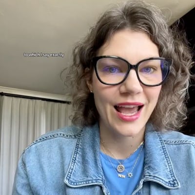 A woman with curly hair and glasses is animatedly speaking, wearing a denim jacket over a blue shirt. She gestures with her hands, engaging the viewer.
