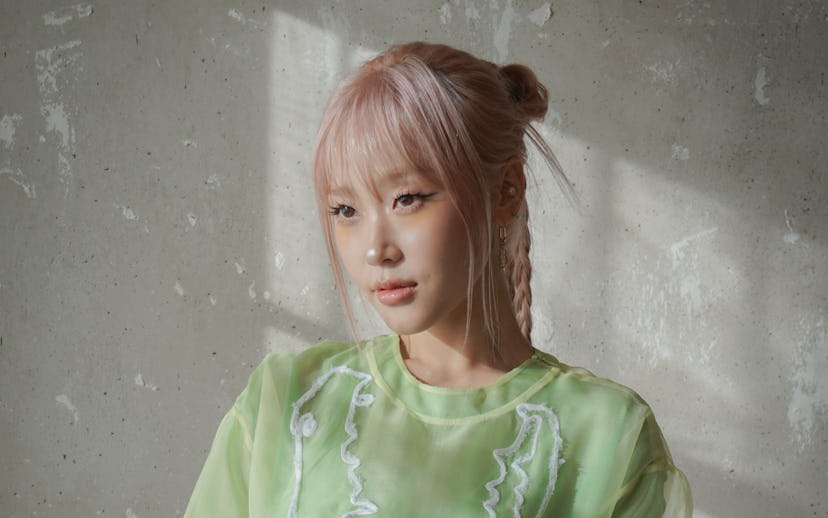 A young person with light pink hair poses thoughtfully in a sheer, green oversized shirt. Soft light...
