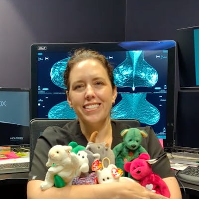 A smiling woman in scrubs sits in an office, holding a collection of colorful stuffed animals. Monitors displaying medical imagery are visible behind her.