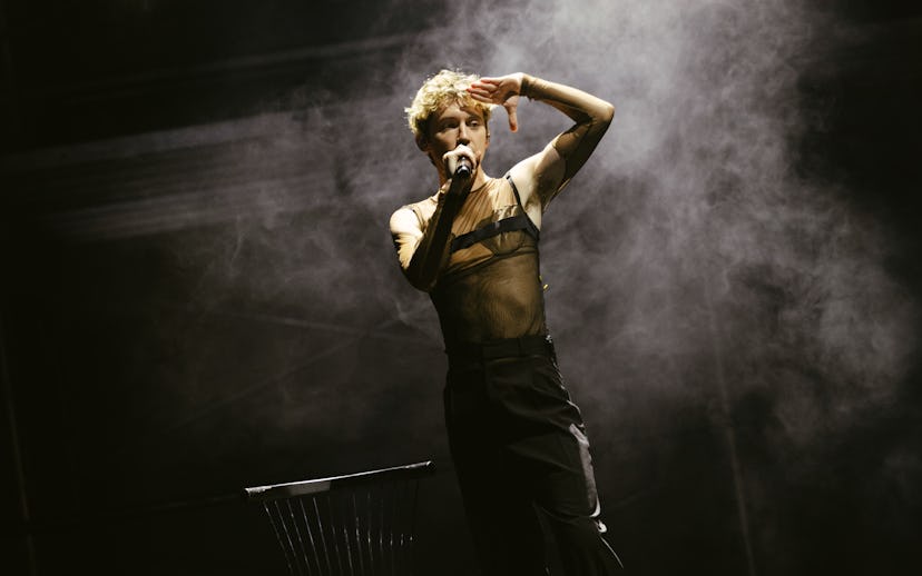 A performer stands on stage, microphone in hand, wearing a sheer top and black pants. Smoke surround...
