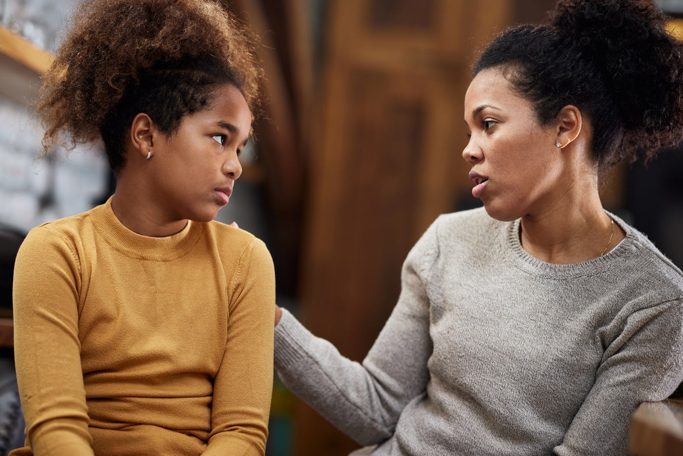 A mother in a gray sweater talks to her daughter, who wears a mustard-colored top. They are engaged in a serious conversation in a cozy setting.