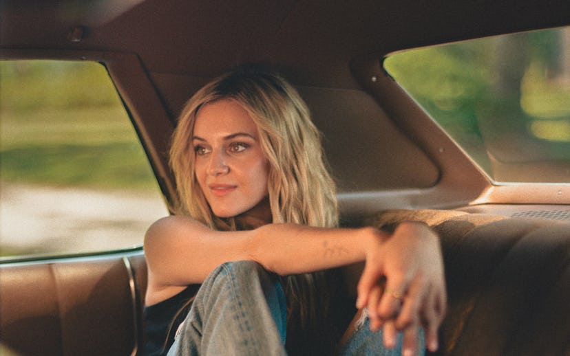 Kelsea Ballerini sits casually in the backseat of a car, wearing a tank top, jeans, and cowboy boots...