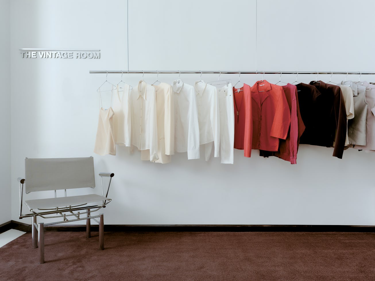 A minimalist room featuring a gray chair and a rack displaying an array of vintage clothing in neutr...