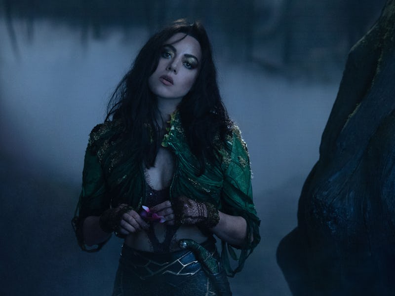 A woman stands in a misty, dark setting, wearing a green, shimmering outfit. Her long dark hair and ...
