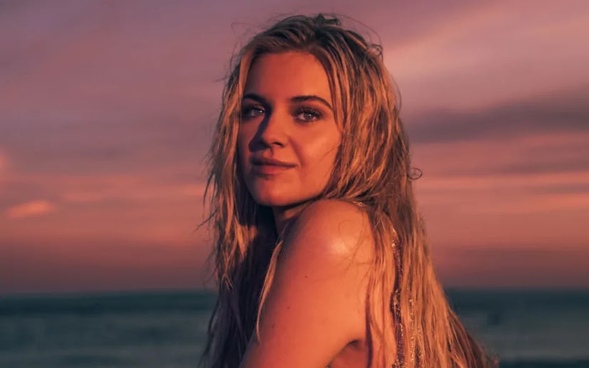 A woman with long, wet hair gazes toward the camera against a vibrant sunset sky, with a serene ocea...