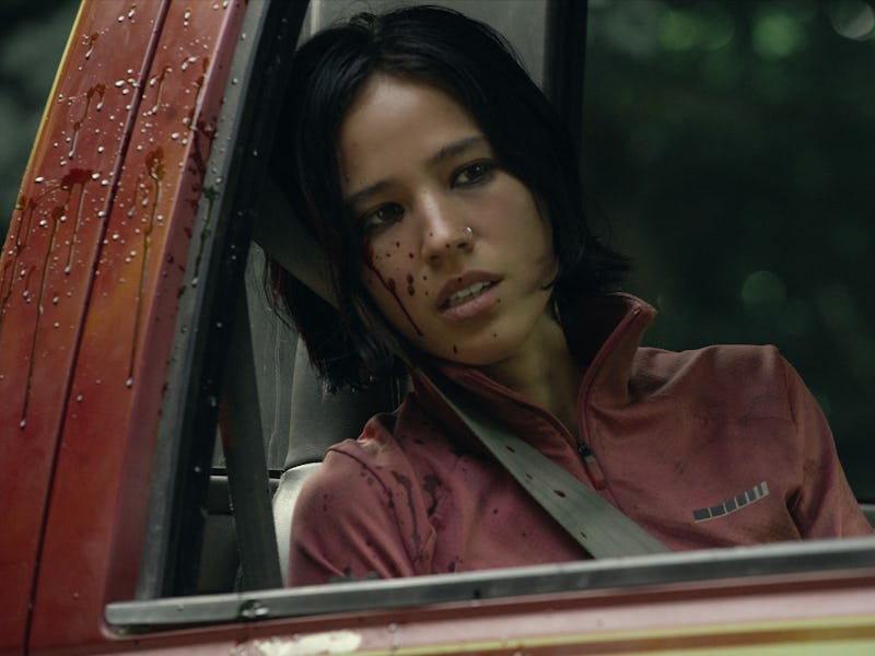 A woman with short dark hair and a nose ring sits inside a reg car splashed with blood.