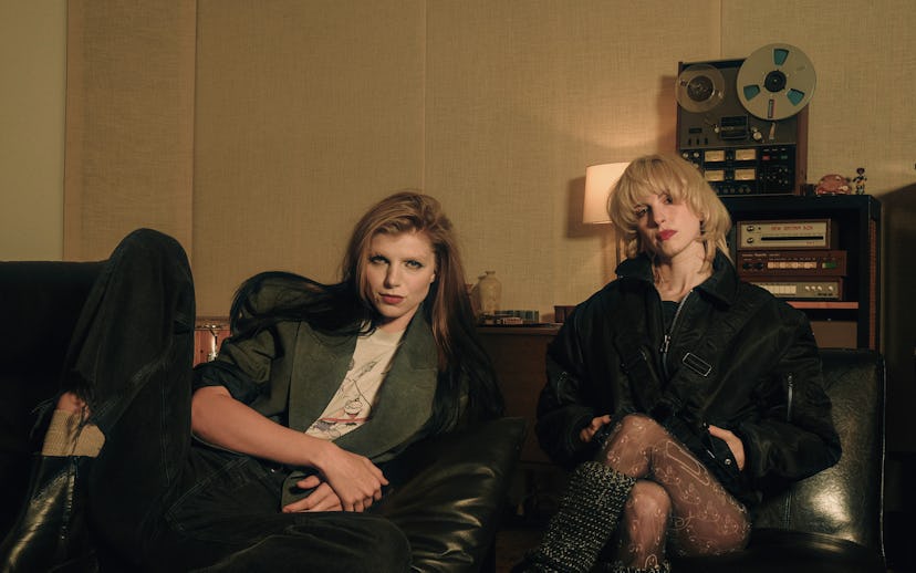 Two young women sit on a leather sofa in a stylish, retro-inspired room, dressed in edgy fashion, wi...