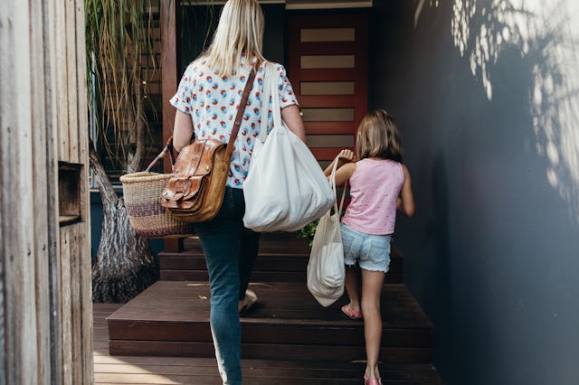 A mom with a crossbody bag and a young girl walk up wooden steps towards a doorway, carrying various...
