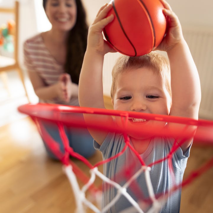 A young child joyfully holds a basketball above a hoop, smiling widely. In the background, an adult ...