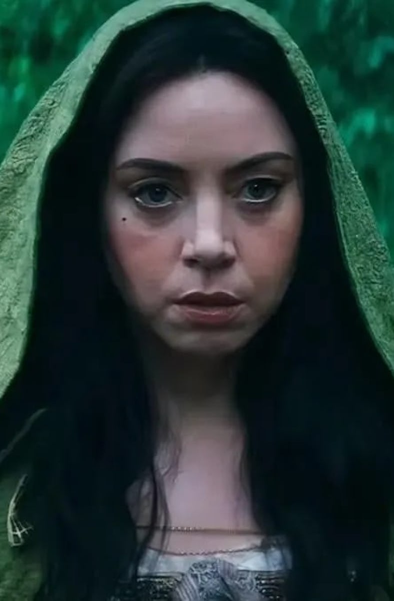 A woman with long dark hair wears a green hooded cloak, standing in a lush, green forest. Her expres...