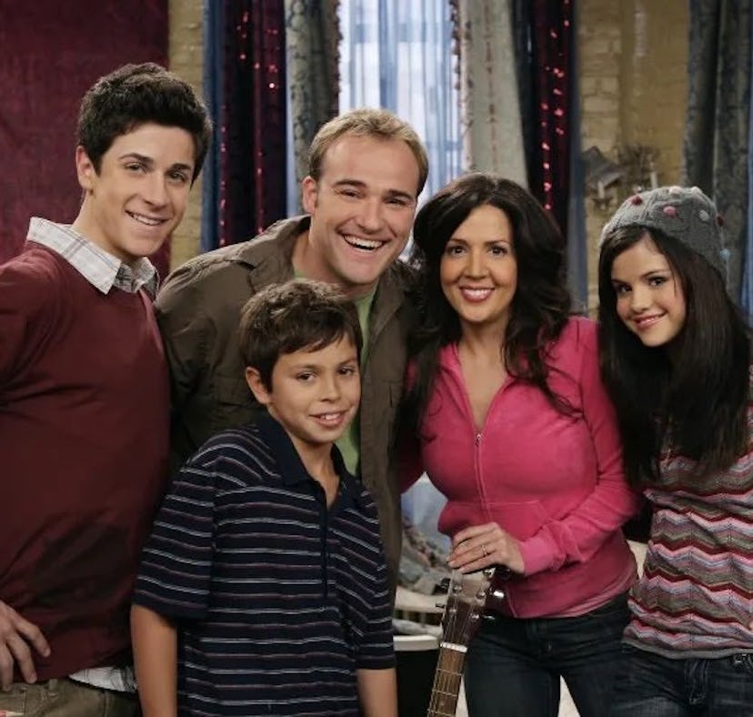 A group of five smiling individuals stands together in a colorful, cozy living room. They appear to ...
