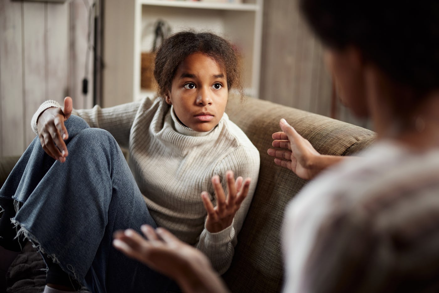 A young girl sitting on a couch looks concerned while engaging in a conversation, gesturing with her hands as she listens intently.