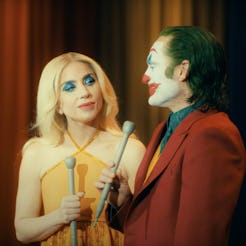 A woman in a yellow dress with bold makeup stands beside a man in a red suit and face paint. They ho...